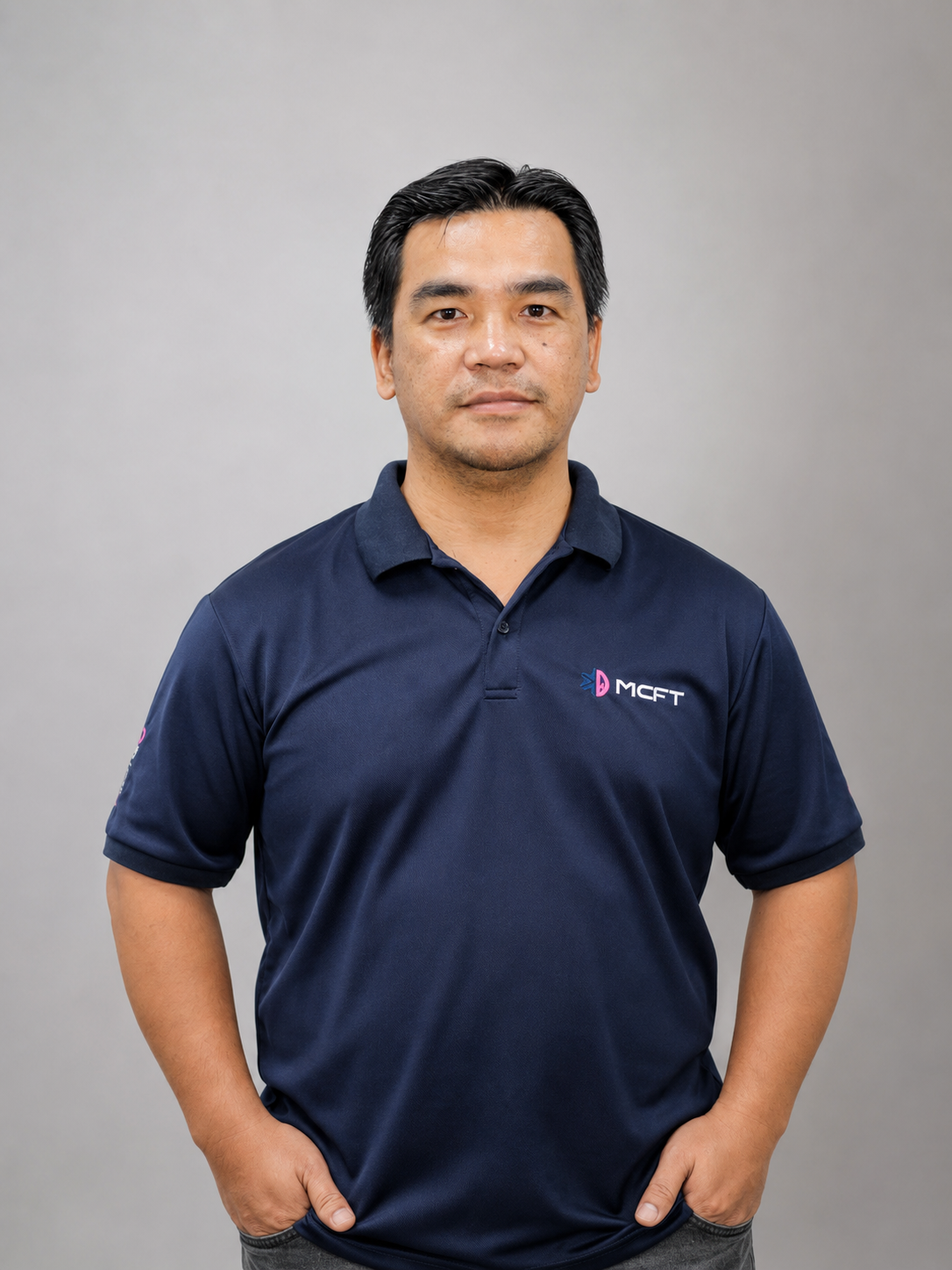 A man standing against a plain gray background, wearing a navy blue polo shirt with a logo that reads 'MCFT'.