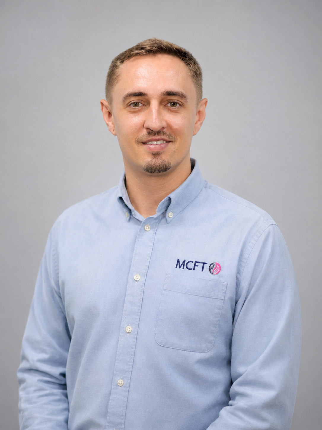 A man with short light brown hair, blue eyes, and light skin, wearing a light blue button-up shirt with a logo on the chest, standing against a plain gray background.