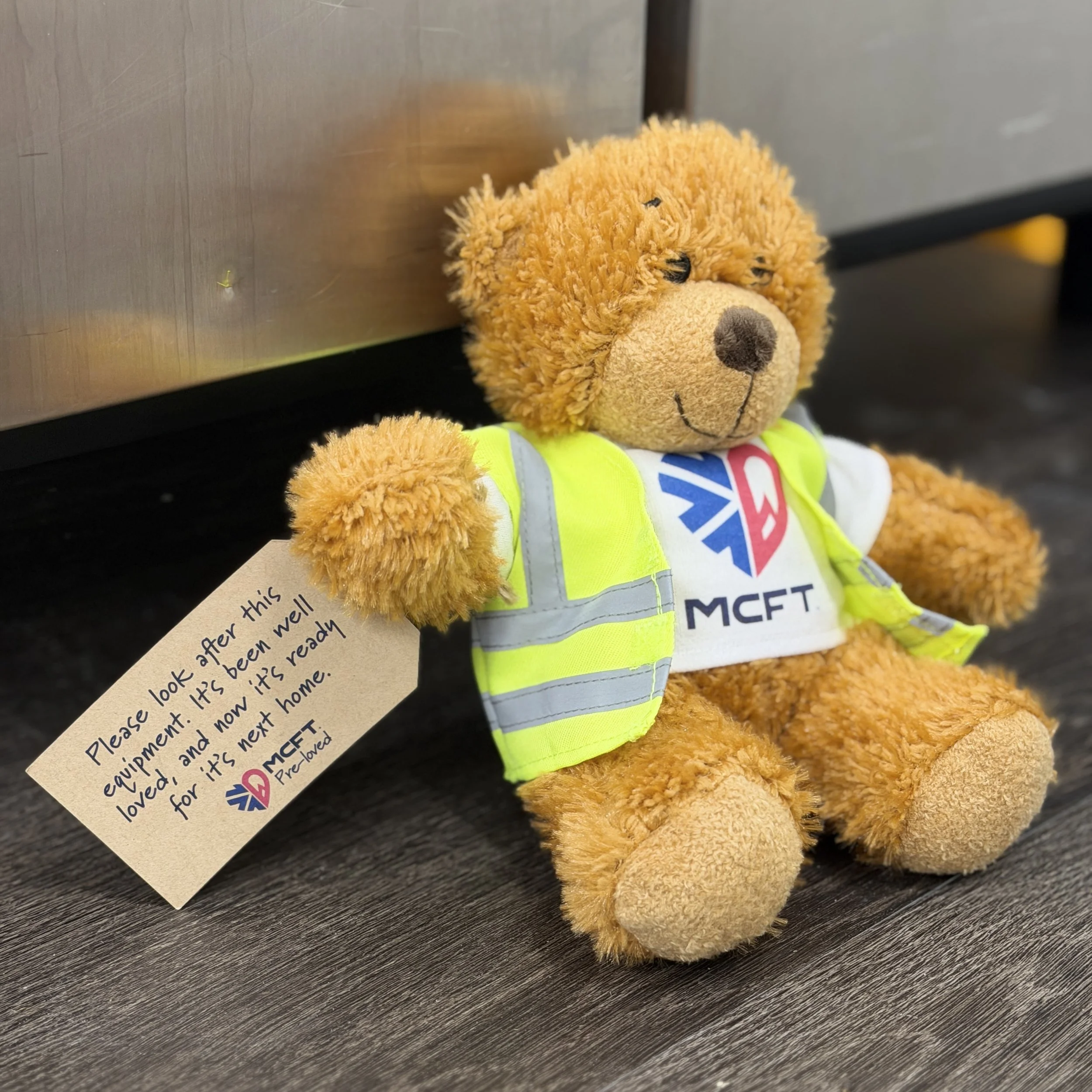 A teddy bear with fuzzy orange fur, wearing a bright yellow high-visibility vest, sitting against a wooden surface, partially obscured by a handwritten note.