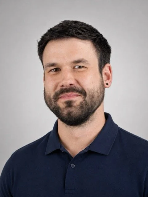 A man with short dark hair and a beard, wearing a dark blue polo shirt, looking into the camera with a slight smile, against a plain gray background.