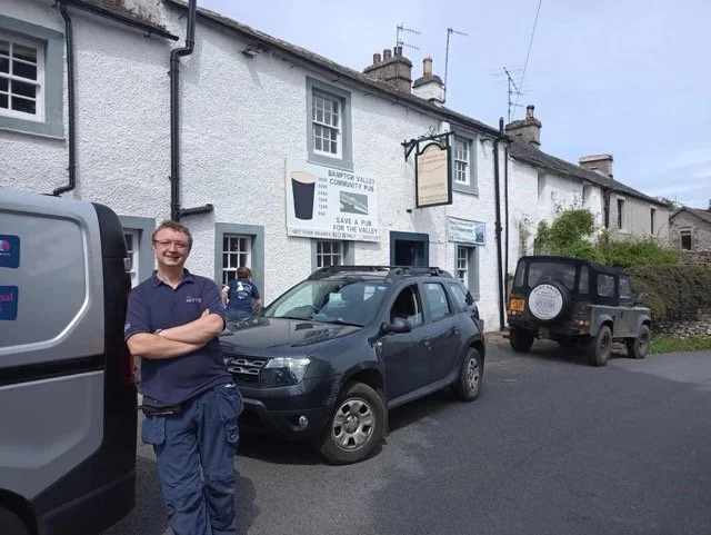 A man in a blue uniform standing with arms crossed outside a pub on a street with cars parked in front. There is a pub sign and a banner advertising a community pub initiative.