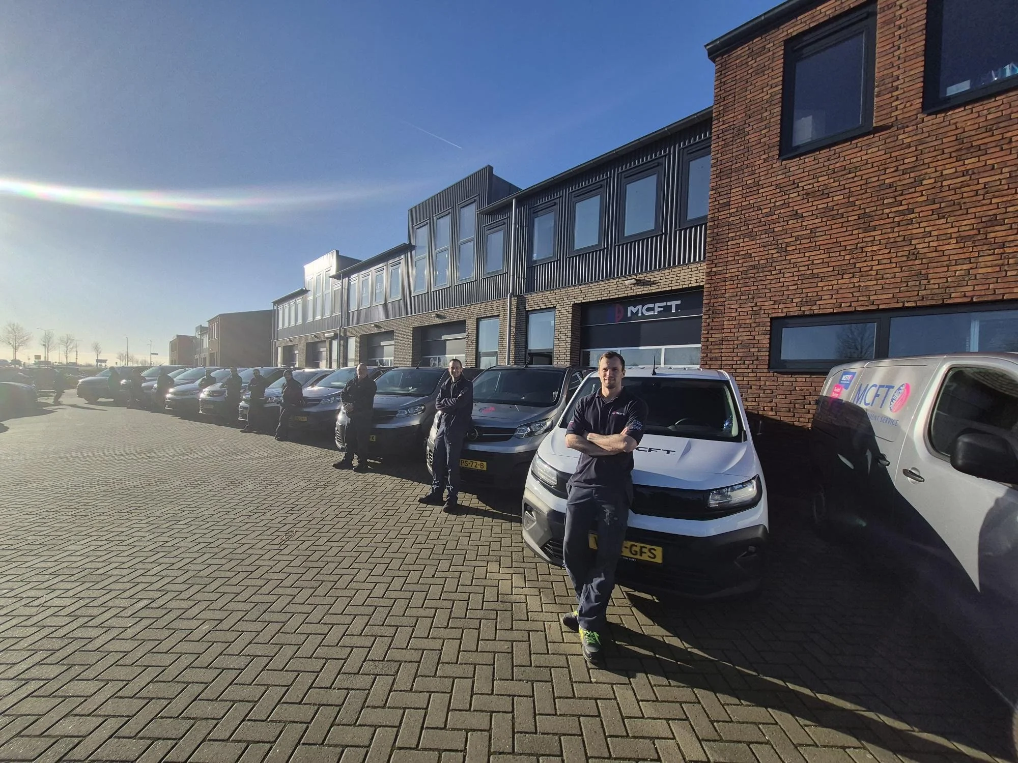 Multiple cars parked in front of a modern commercial building with large windows and brick and black metal exterior walls. Several people stand alongside the cars, with a man in the foreground crossing his arms and smiling. The sky is clear with a visible contrail.