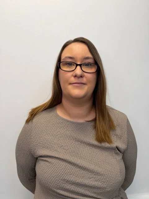 A woman with glasses and long brown hair standing against a plain white wall.