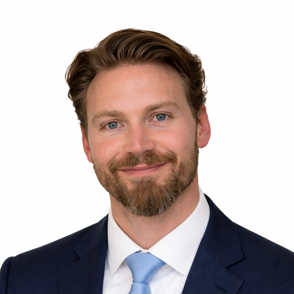 Portrait of a man with light brown hair, blue eyes, and a beard, wearing a navy blue suit, white shirt, and light blue tie, smiling against a white background.