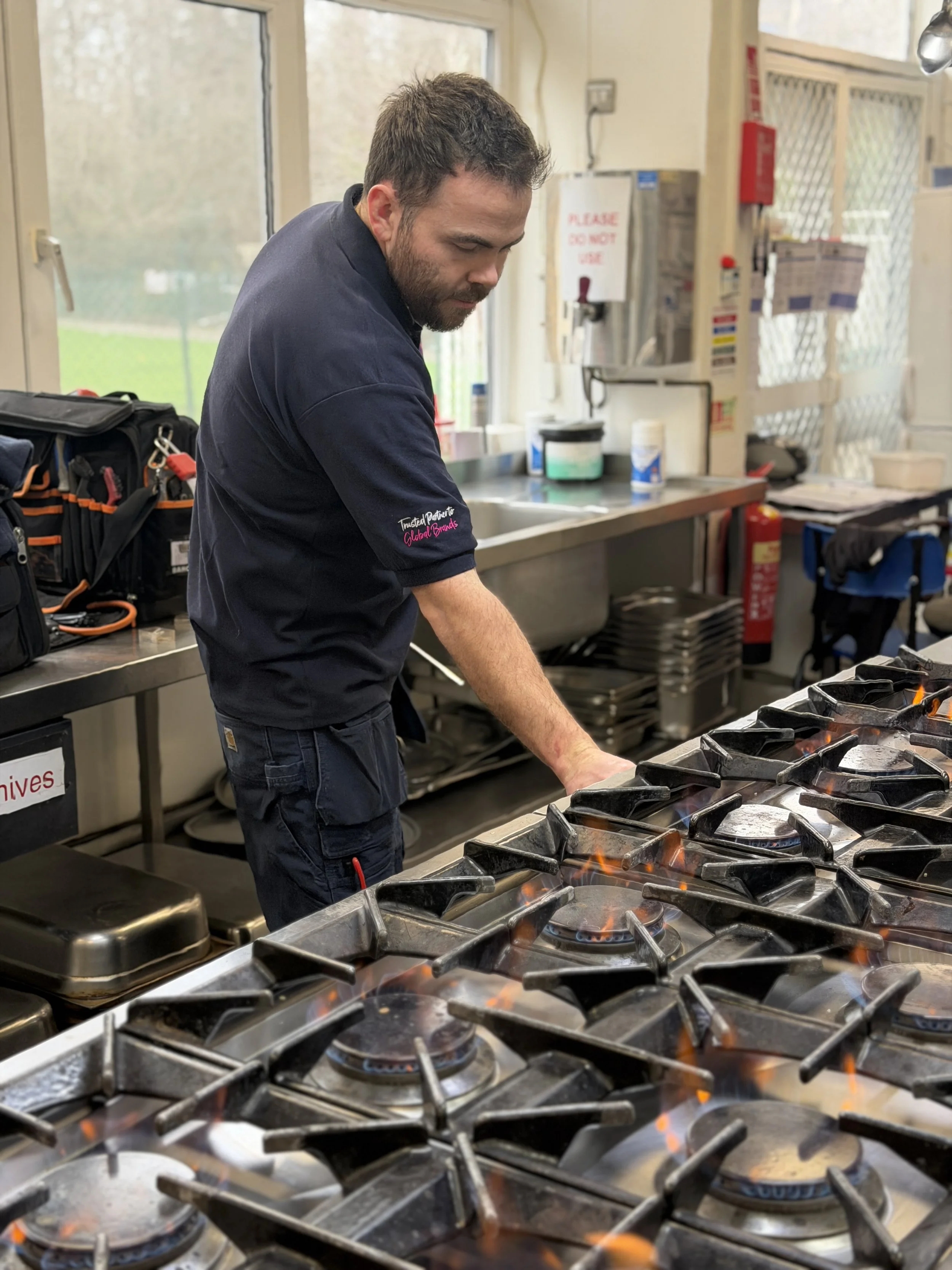 A man with a beard, wearing a black shirt, standing at a commercial kitchen stove, cooking over open flames with pots and kitchen equipment visible in the background.