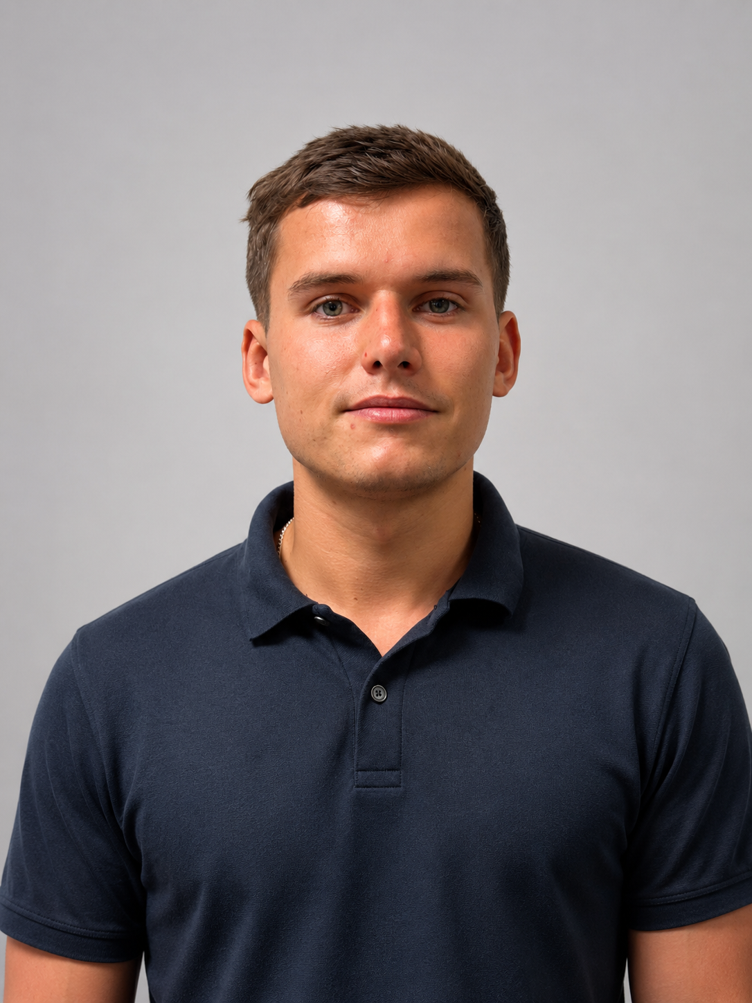 Portrait of a young man with short brown hair wearing a dark polo shirt standing against a gray background.