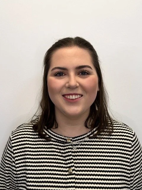 A young woman with shoulder-length brown hair, wearing a black and white striped shirt, smiling against a plain white wall.