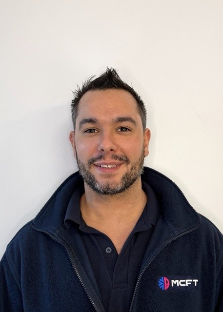 Man with short dark hair and a beard wearing a black polo shirt and a dark zip-up jacket with a company logo, standing against a plain white background.