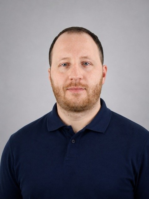 Portrait of a man with short brown hair, beard, wearing a navy blue polo shirt, against a gray background.