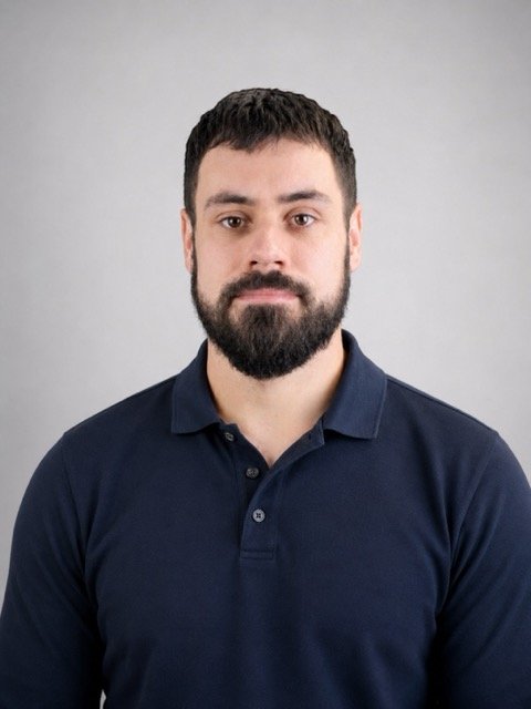 A man with dark hair and a beard wearing a navy blue polo shirt looks directly at the camera against a plain gray background.