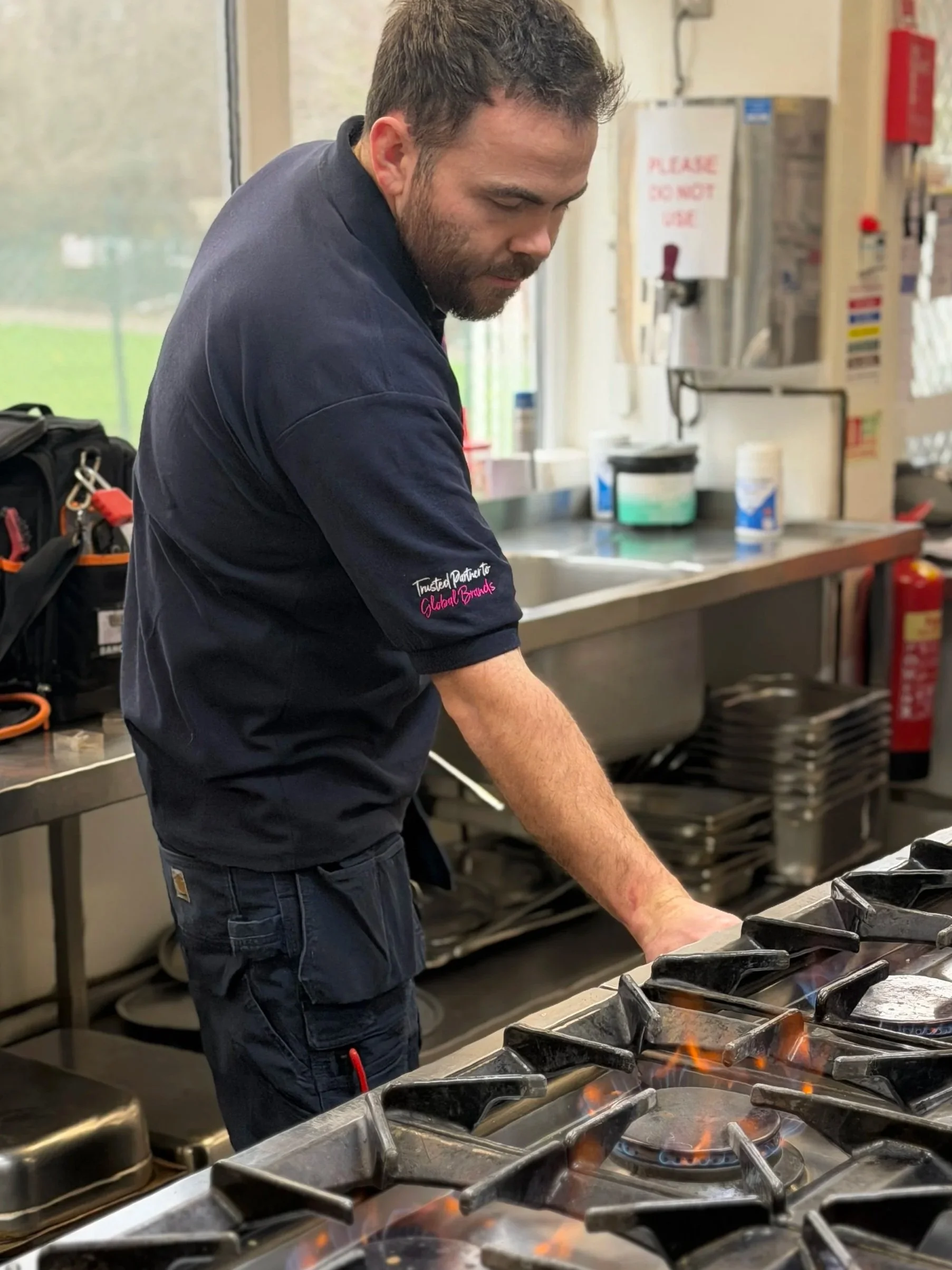 A man working in a commercial kitchen, standing near a stove with flames, with kitchen equipment and containers around him.
