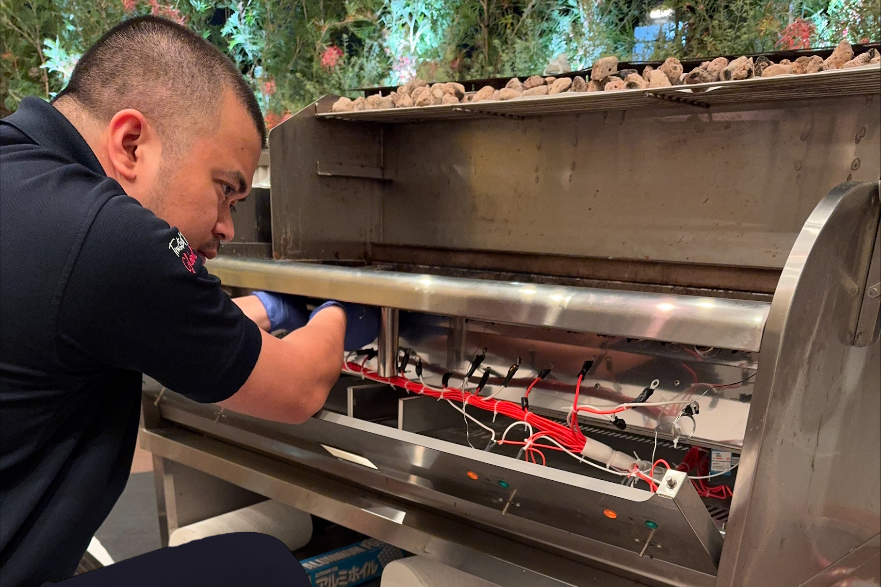 A technician working on an open electronic machine with circuit wiring and components, its outer casing removed, in an indoor setting with greenery in the background.