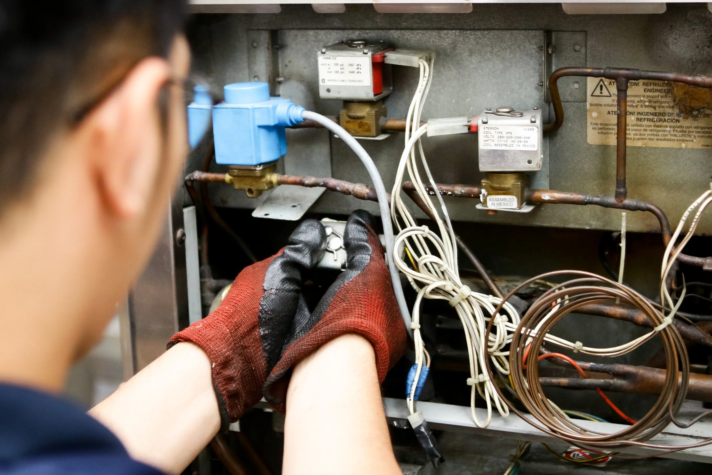 Person wearing gloves working on electrical components inside an open panel, with various wires, switches, and labels visible.