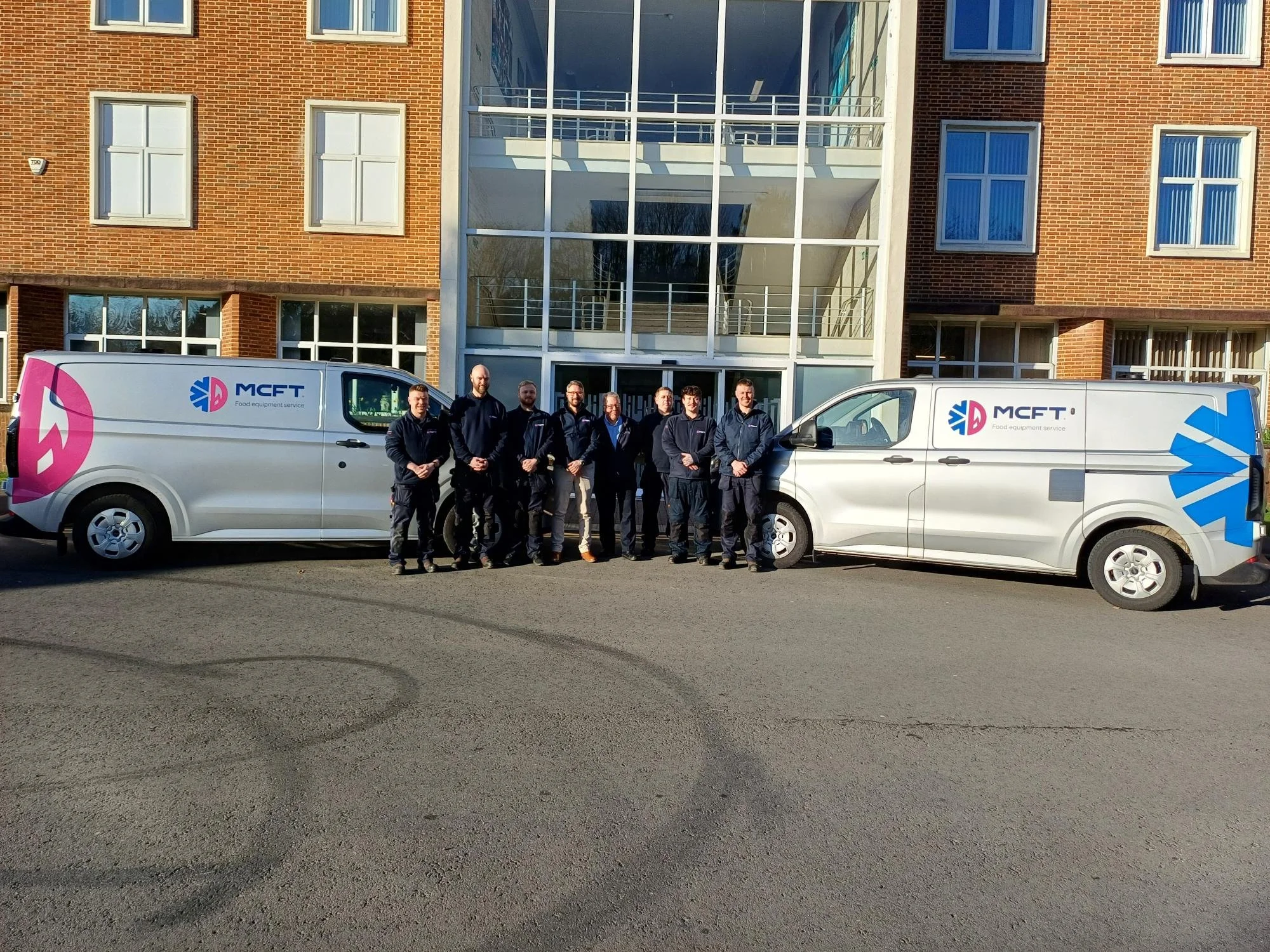 Group of seven people standing in front of two white company vans with the logo "MCFT" parked on a paved street outside a building with brick walls and large glass windows.