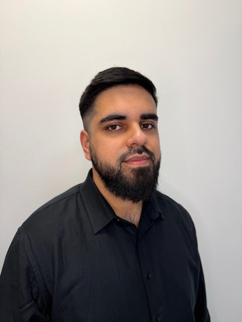 A man with dark hair and a beard wearing a black button-up shirt stands against a plain white wall.