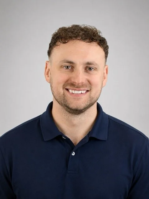 Headshot of a smiling man with short curly hair, wearing a navy blue polo shirt against a plain gray background.