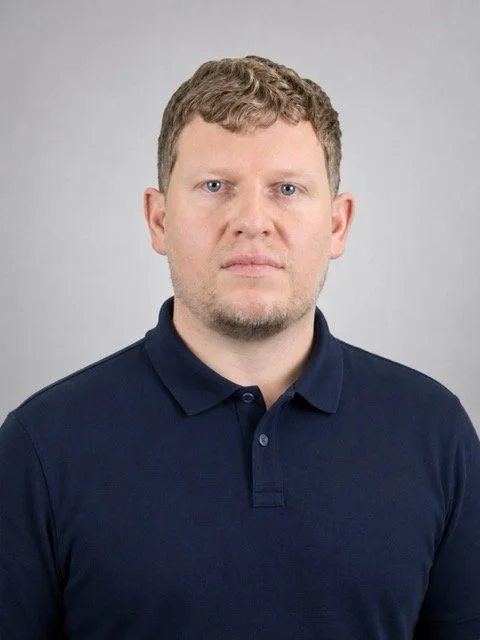 A man with short, curly light brown hair and a serious expression, wearing a navy blue polo shirt, standing against a plain light gray background.