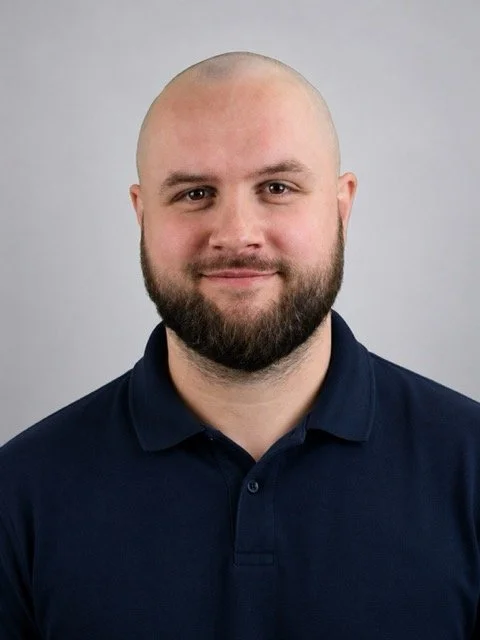 Portrait of a man with a shaved head and a full beard, wearing a navy polo shirt, smiling at the camera against a plain gray background.