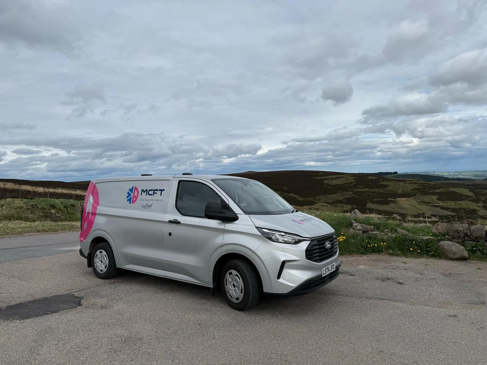 White delivery van with 'MCET' logo parked on a rural road with rolling hills and cloudy sky in the background.