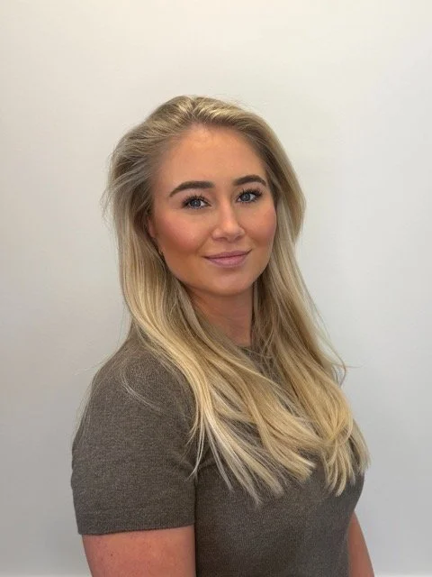 Portrait of a young blonde woman with long hair standing against a plain white wall, wearing a gray top.