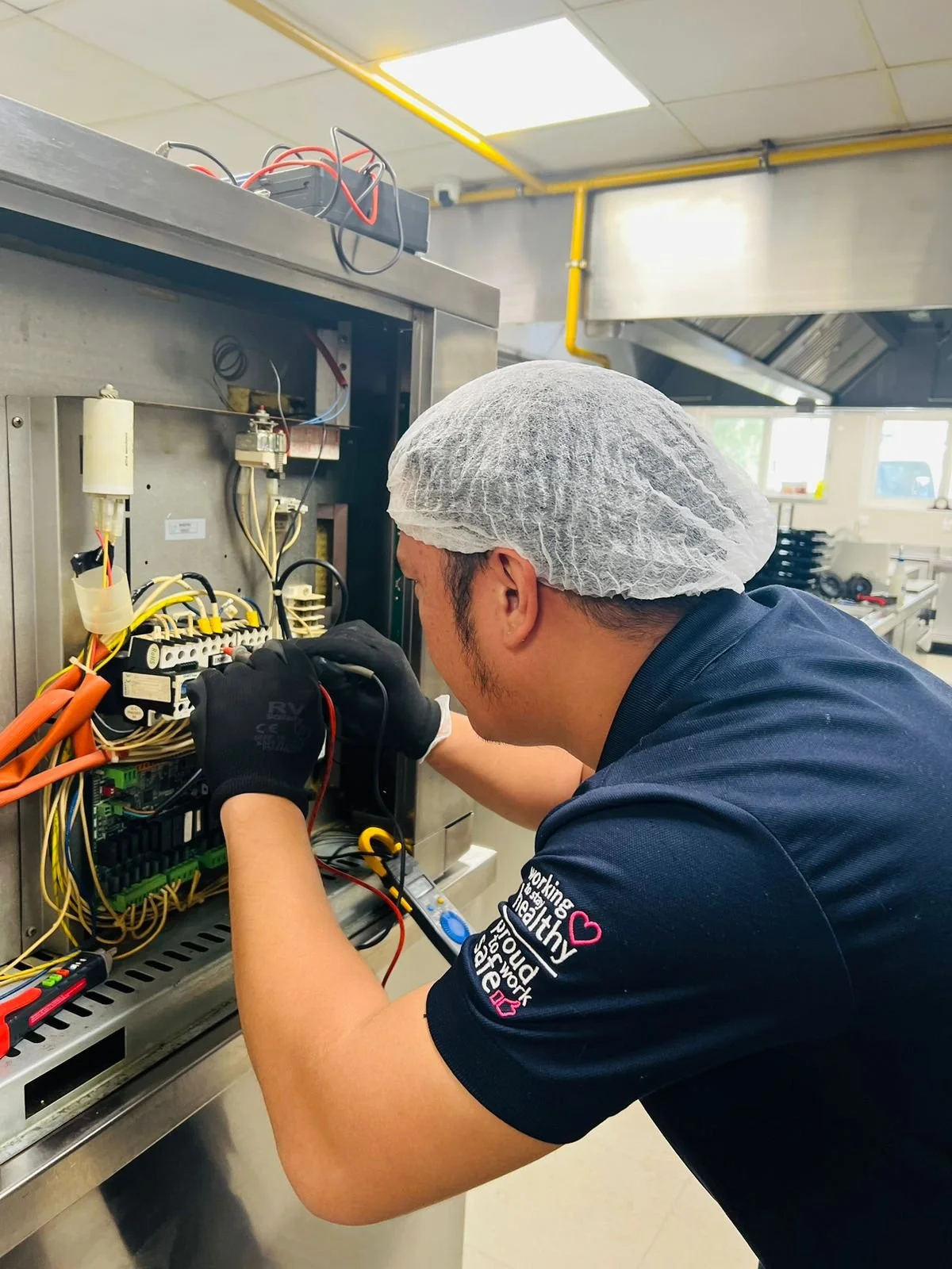 A technician wearing a hair net and gloves working on electrical wiring inside an industrial control panel in a factory setting.