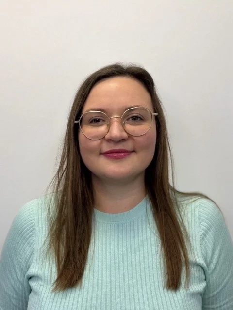 A woman with glasses and long brown hair, wearing a light blue sweater, posing against a plain white wall.