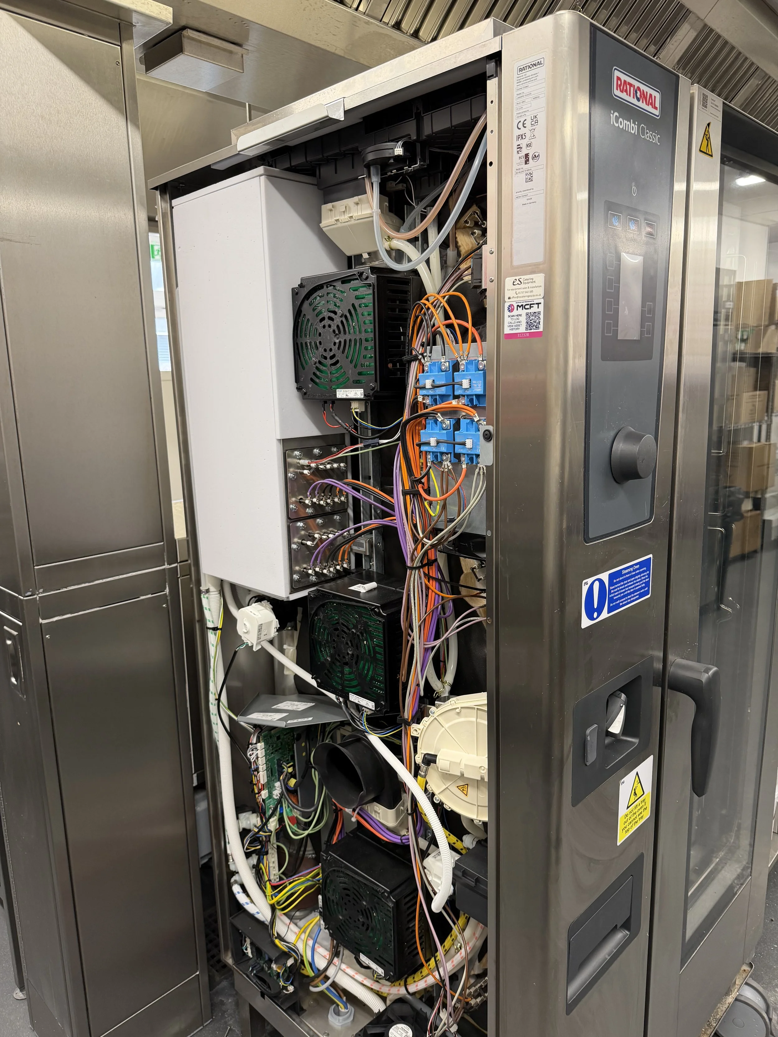 An open stainless steel kitchen appliance with internal wiring, circuit boards, fans, and various electronic components.