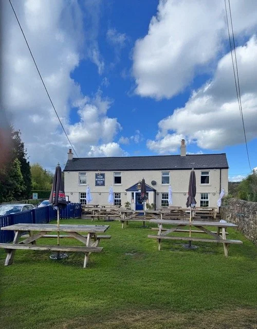 Exterior of a pub with outdoor seating, picnic tables, umbrellas, under a partly cloudy sky.