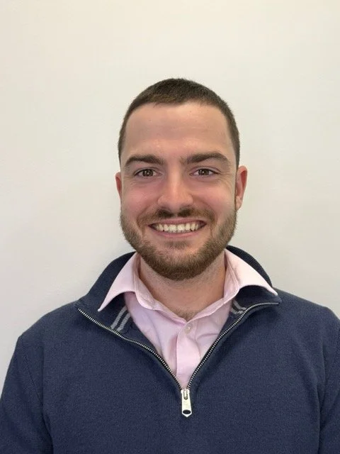 A smiling man with short dark hair and a beard, wearing a pink collared shirt and a navy zip-up jacket, standing against a plain white wall.