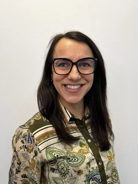 Woman with long dark hair, wearing glasses and a patterned shirt, smiling in front of a plain white wall.