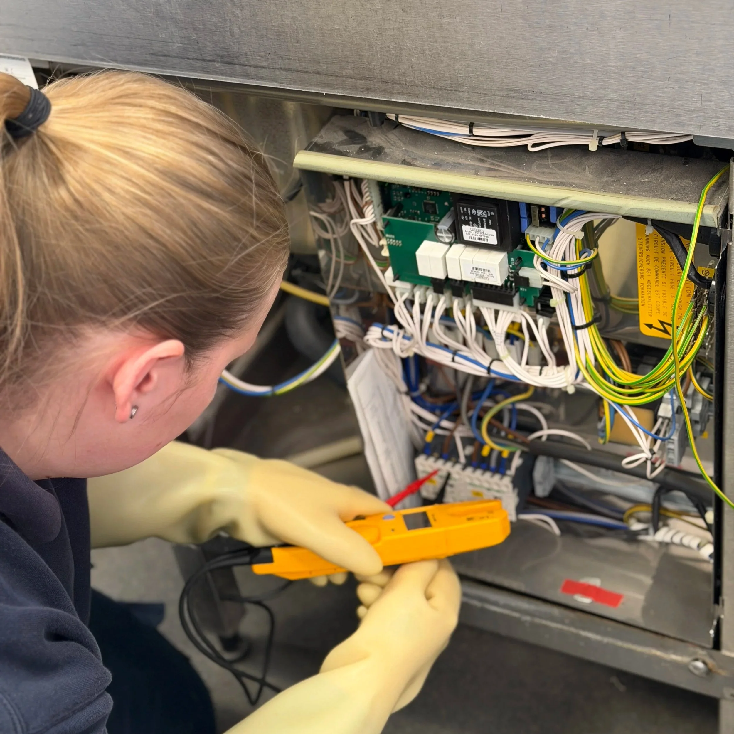 Close-up of a person with blonde hair working on electrical panels filled with numerous wires and circuit boards.