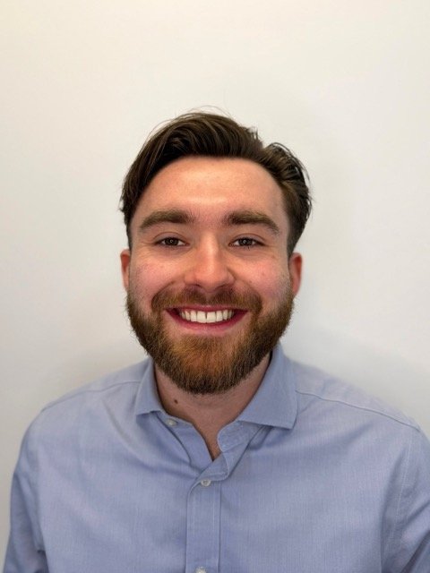 A smiling man with dark hair and a beard, wearing a light blue button-up shirt against a plain white background.