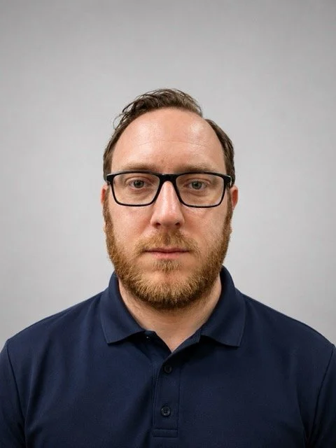 A man with glasses, a beard, and short hair wearing a navy blue collared shirt, looking directly at the camera against a plain background.