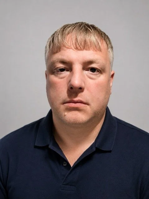 A close-up portrait of a man with short blonde hair and a serious expression, wearing a dark collared shirt, against a plain background.