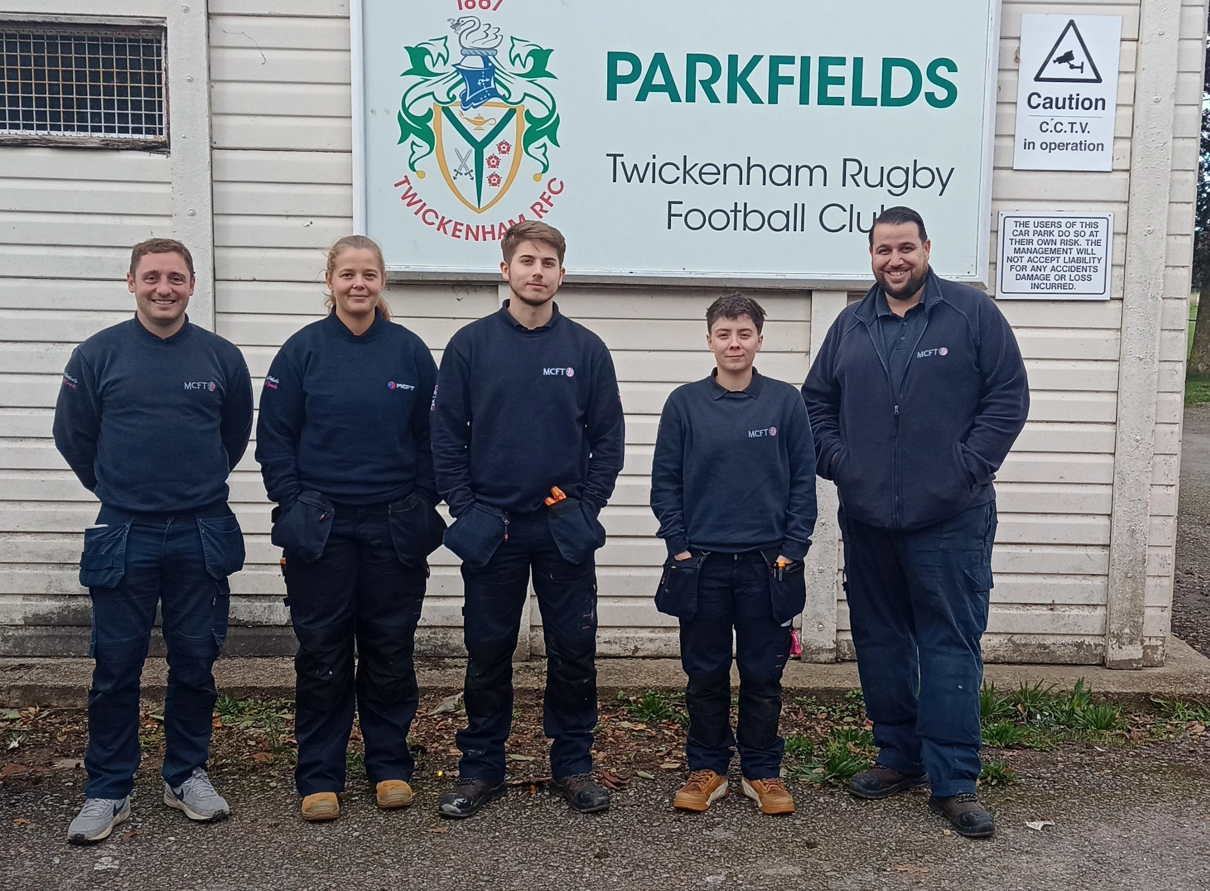 Five people standing in front of a sign that reads 'Parkfields Twickenham Rugby Football Club.' The people are wearing dark blue jackets with 'MCFT' and a logo, smiling, and looking at the camera.