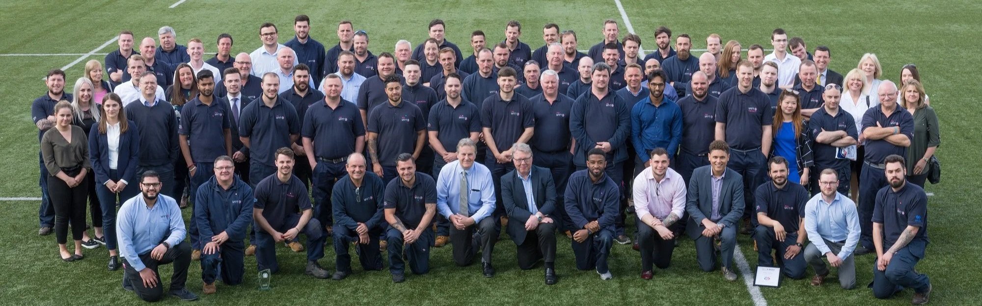 Group photo of many men and women on a football field, some wearing dark blue polo shirts with a logo, others in business casual attire, posing for a team or company picture.