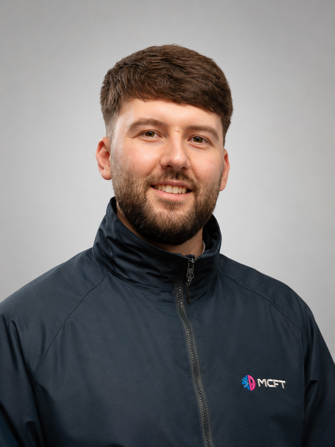 Headshot of smiling young man with brown hair and beard, wearing a black zip-up jacket with a logo on the chest, against a gray background.