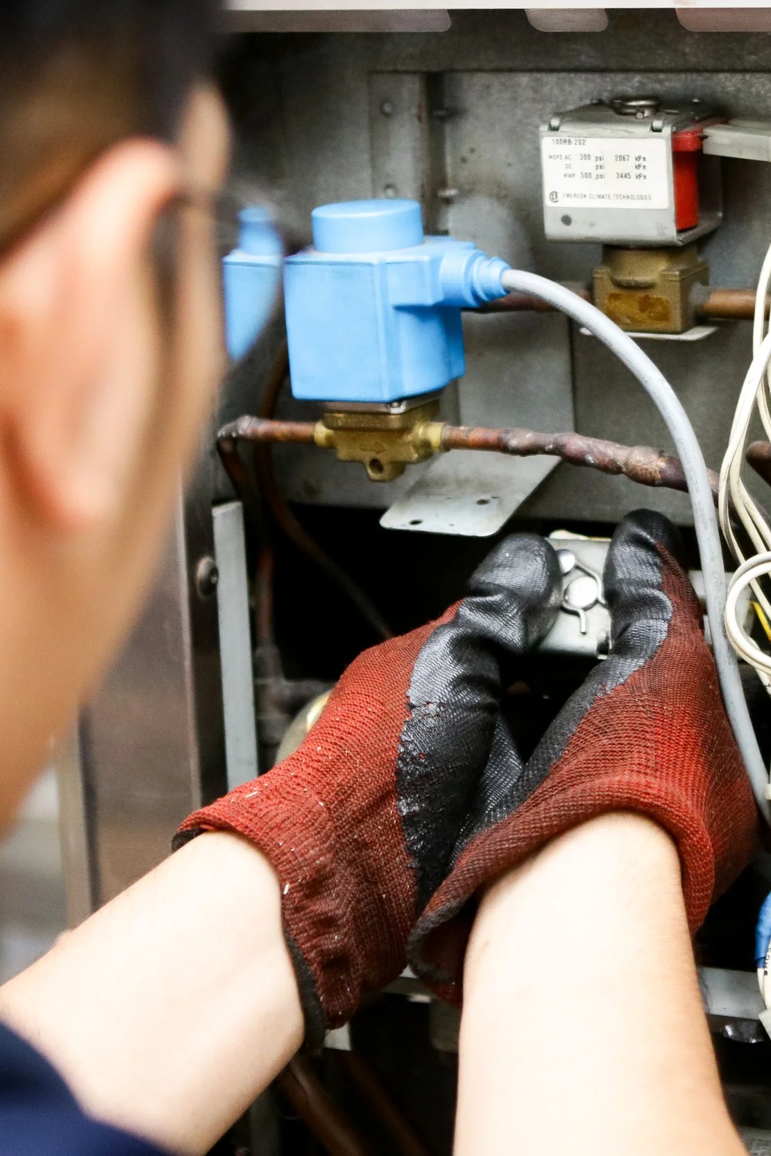 A person wearing red work gloves working on an electrical panel or appliance, with their hands near wires and components inside the panel.