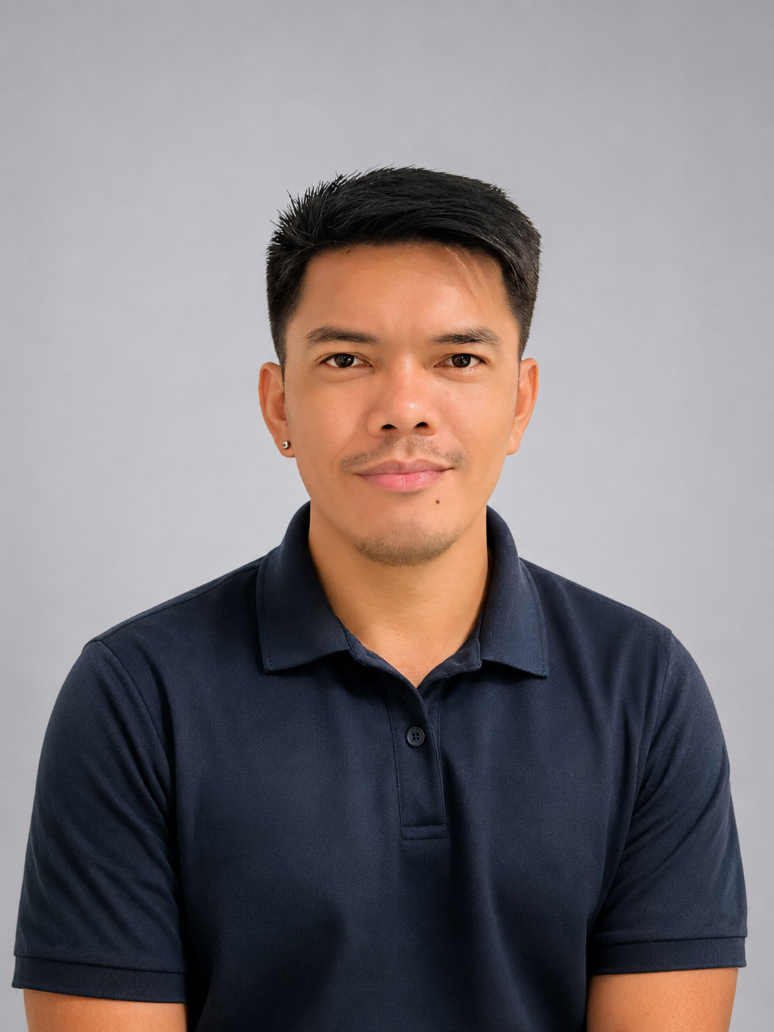 A young man with short black hair, wearing a navy polo shirt, looking at the camera with a slight smile, against a plain gray background.
