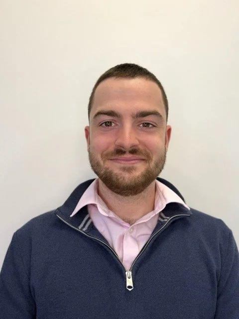 A young man with short brown hair and a beard, wearing a pink collared shirt and a navy blue zip-up jacket, smiling in front of a plain white wall.