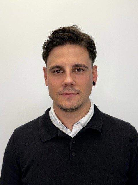 Portrait of a young man with short dark hair, wearing a white collared shirt and a black sweater, standing against a plain white background.