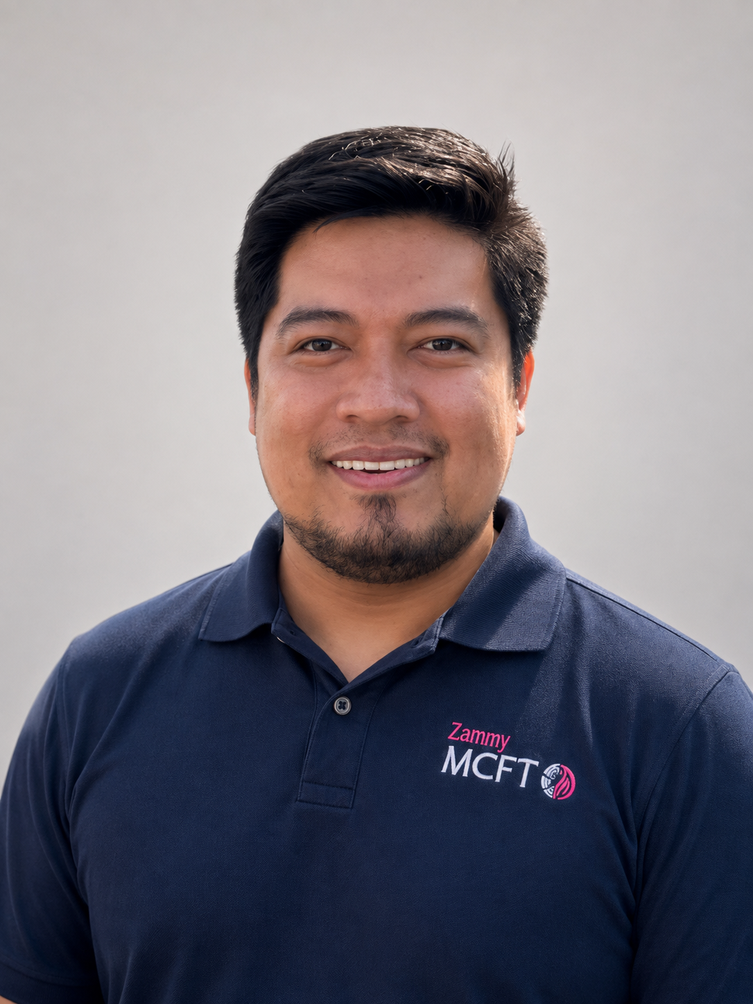 A man with short dark hair, a beard, and a mustache, wearing a navy blue polo shirt with 'Zammy MCFT' embroidered on it, smiling at the camera, standing against a plain light gray background.