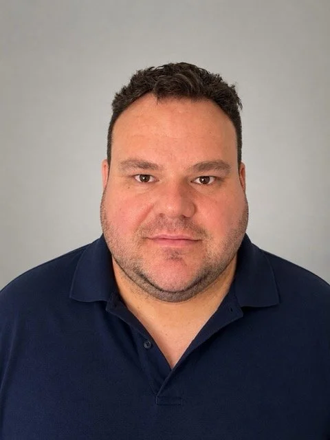 A man with short dark curly hair, wearing a navy blue collared shirt, looking directly at the camera, against a plain light gray background.