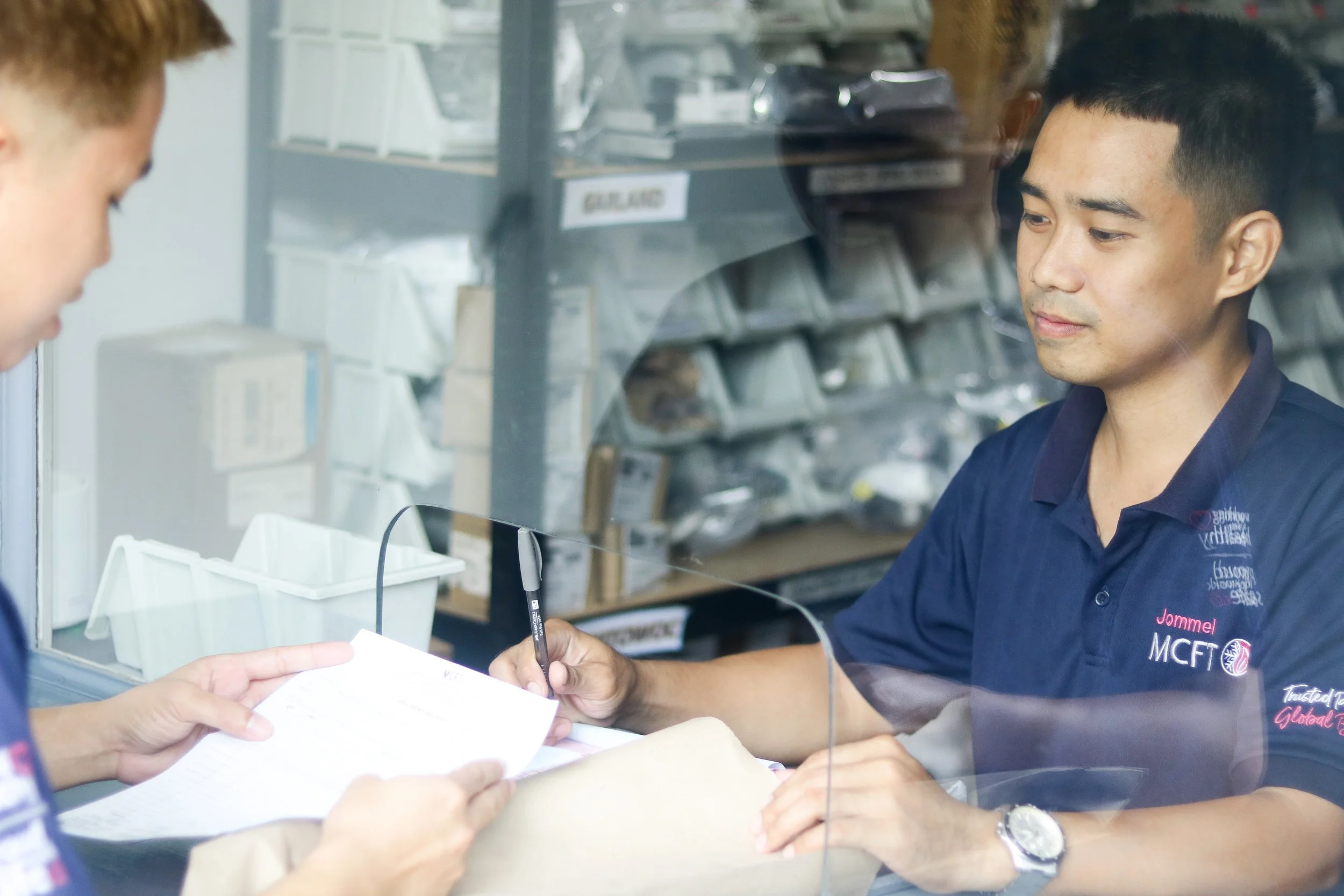 A man in a blue uniform is sitting behind a glass window, working with a woman across from him at a counter. The man is holding a pen and appears to be filling out or reviewing paperwork. In the background, there are shelves with various items and a sign that reads 'GEAR.'