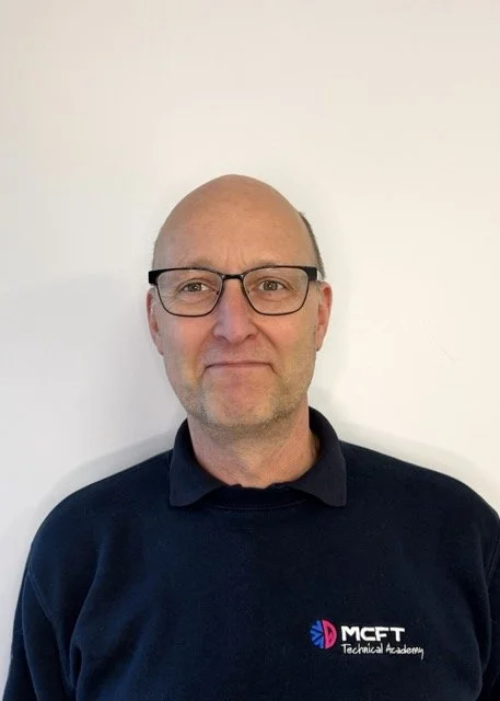 A middle-aged man with glasses and a bald head wearing a dark sweater with the MCFT Technical Academy logo, standing against a plain white wall.