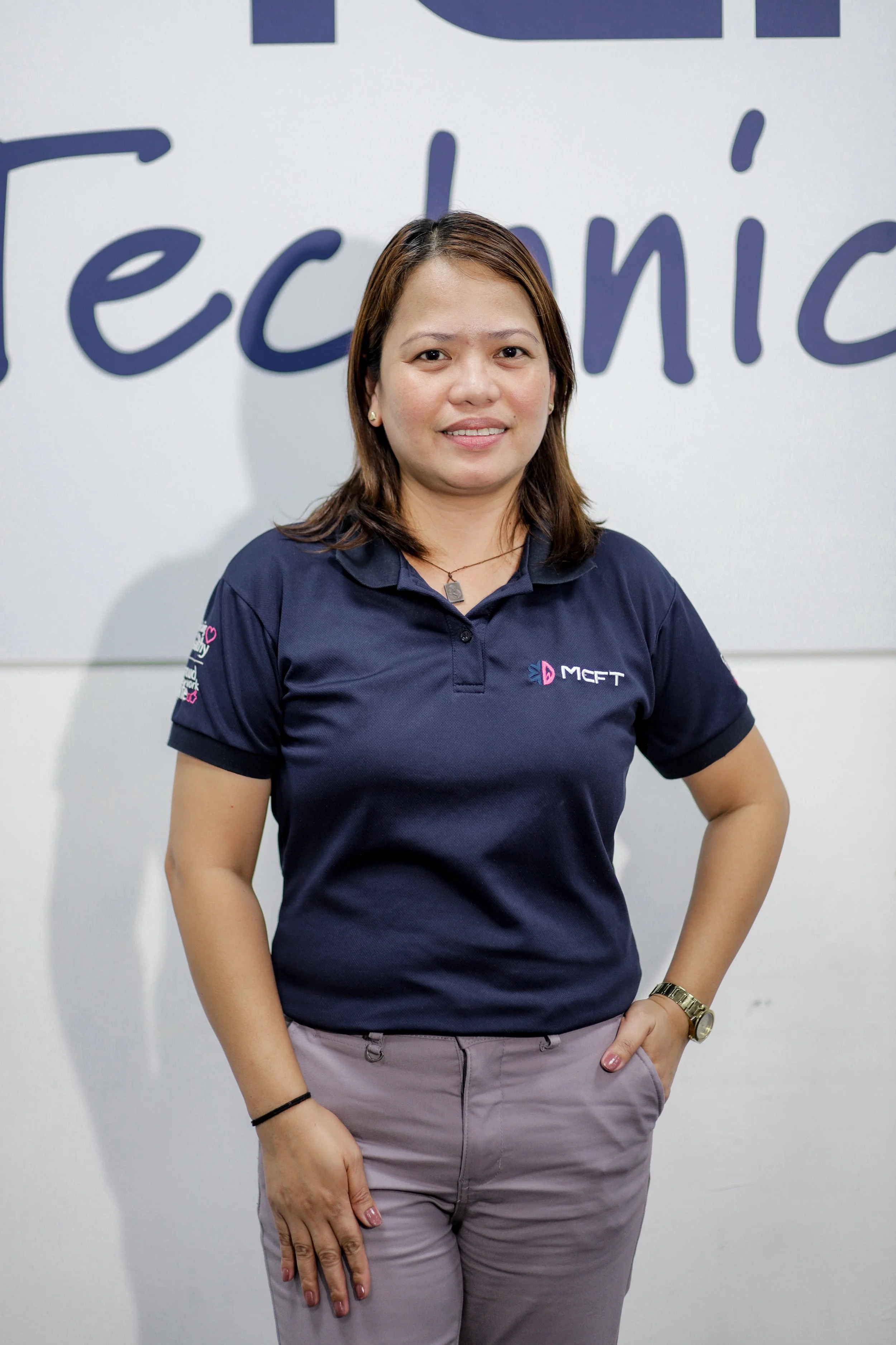 A woman standing in front of a white wall with colorful text design, wearing a navy blue collared shirt with logo and text, beige pants, a watch, and jewelry, smiling at the camera.