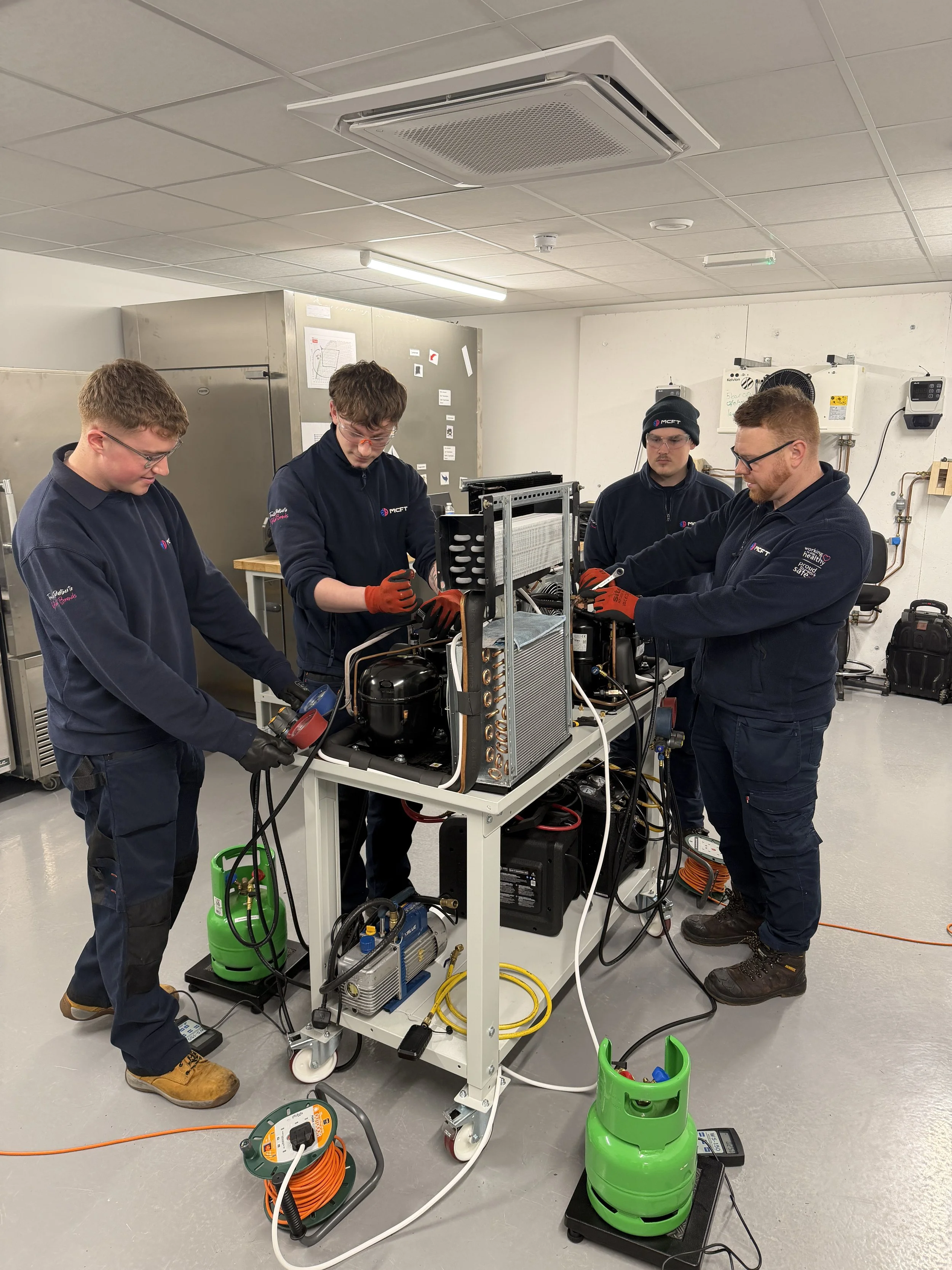 Four young men working together on a piece of equipment in a laboratory or workshop setting. They are wearing dark uniforms and safety glasses. There are various tools, wires, and equipment around them, and they appear to be engaged in a technical task or experiment.
