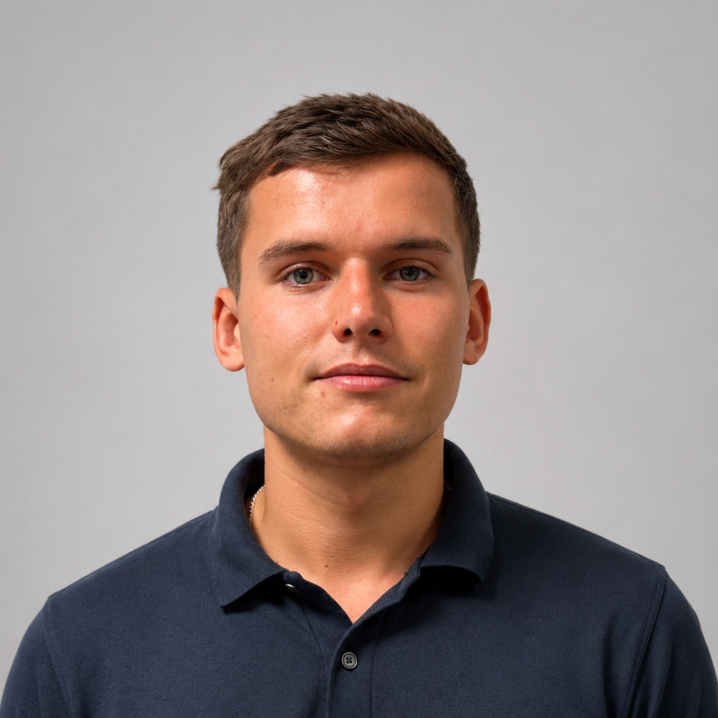 Portrait of a young man with short brown hair, wearing a dark collared shirt, standing against a plain gray background.