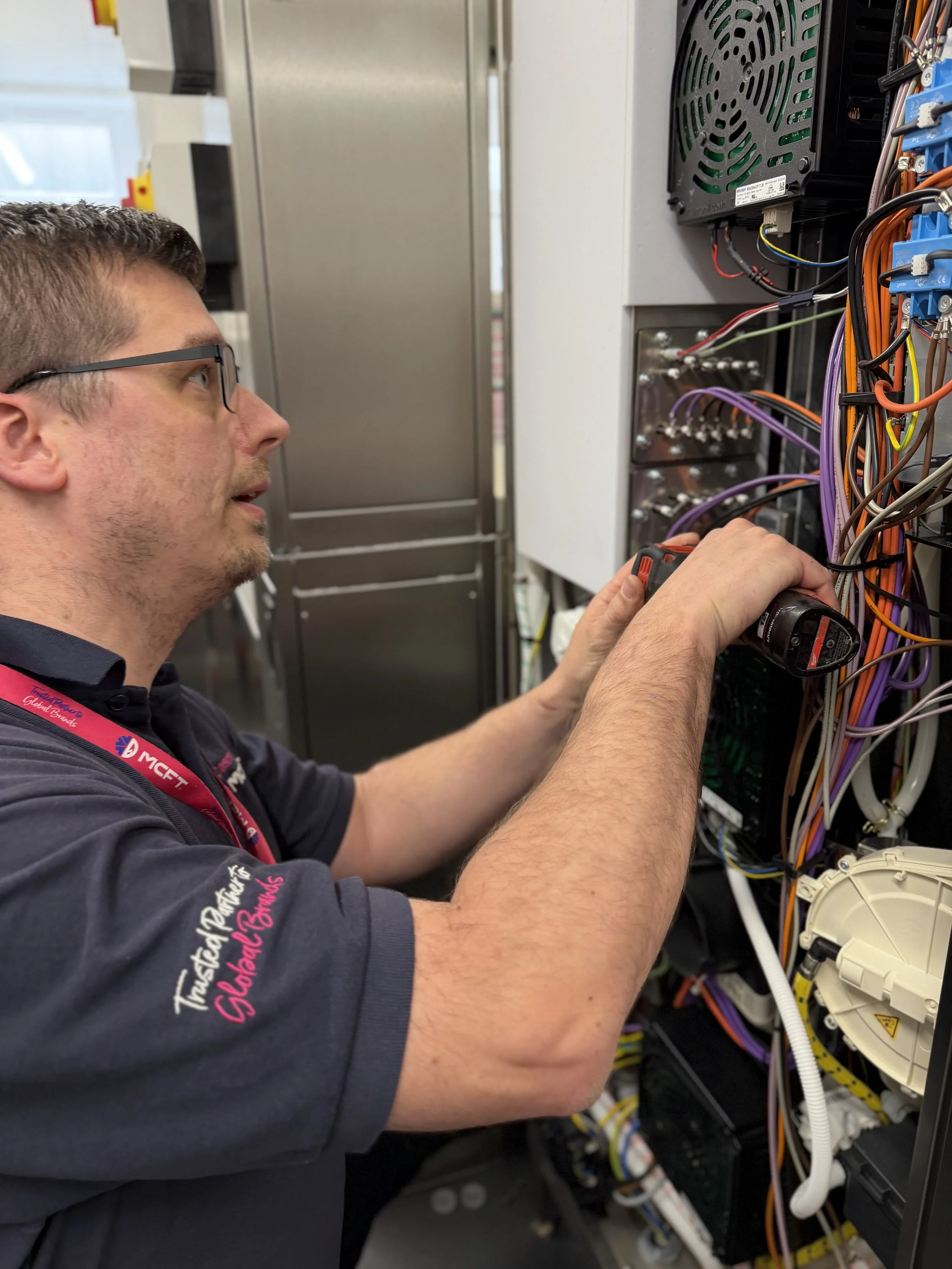 A technician wearing safety glasses and a black shirt working on an electrical panel with multiple colorful wires and circuit components.