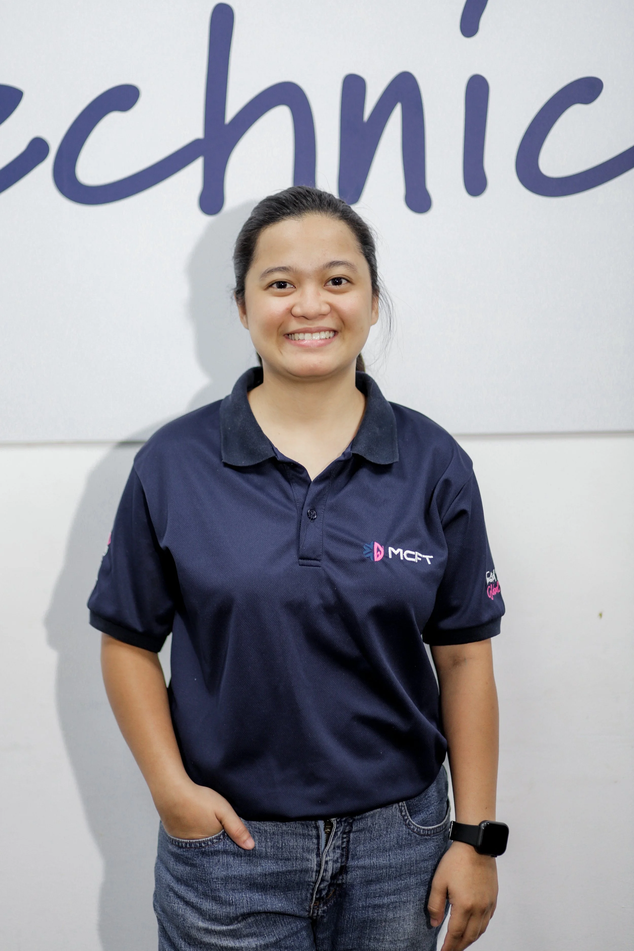 A young woman is standing in front of a white wall with the word 'techie' written on it. She is smiling, wearing a navy blue polo shirt with the 'MCFT' logo, and has her hair pulled back. She's dressed in casual jeans and has a smartwatch on her left wrist.
